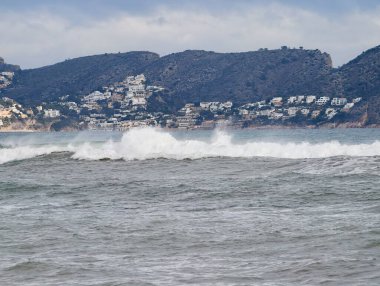 Kıyı yamacına çarpan dalgalı deniz, beyaz sörf hatları, teraslı yamaçlardaki uzak tatil evleri.,