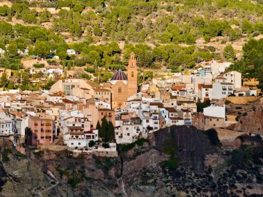 Hava manzaralı uçurum kenarı köyü, gün batımında taş evler engebeli kayalıklara tutunuyor, kilise kulesi dar duvarların üzerinde yükseliyor.