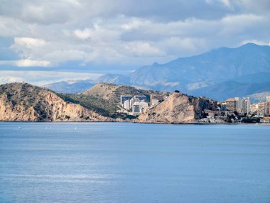 Kıyı kasabası kayalık promontory ve dağ zeminiyle çevrelenmiş, öğlen ışığı, kümelenmiş binalar ve alçak