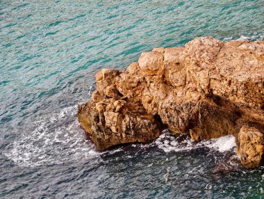 Turkuaz su engebeli kayalara çarpıyor, köpüklü kenarlı canlı renk kontrastı, tenha koy havası.,
