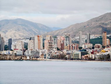 Yüksek kuleleri ve dağ arkaplanı olan kıyı silüeti, sakin deniz önplanı, kümelenmiş binalar