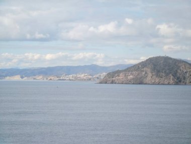 Dağlık arkaplanı ve sakin körfez suları olan uzak bir sahil kasabası. Ufuk çizgisi ve katmanlı tepeler sunuyor.