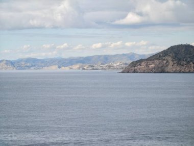 Sakin okyanus uzak kıyı şeridi ve yuvarlanan tepelerle genişliyor, bulutlar yumuşak bulutlarla kaplı, sessiz mavi