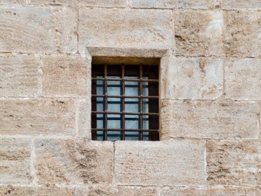 Taş duvara yerleştirilmiş küçük parmaklıklı pencere, dar duvar eklemleri ve yıpranmış yüzey ile küçük kare açıklığı
