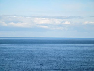 Mavi gökyüzünün altında engin bir açık okyanus, yumuşak dalgalar ve uzak bulut katmanları huzurlu bir ortam yaratıyor, ideal.