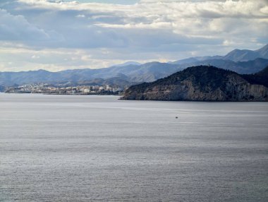 Uzak tepelerle çevrili kıyı kasabası, sakin körfez suları yumuşak bulutları yansıtıyor, kentsel kıyı şeridi birleşiyor