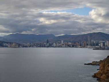 Düşünceli bulut örtüsü altında sakin koy, yumuşak yansımalı parlak su, uzak ufuk çizgisi ve dağ sırtı,