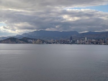 Geniş kıyı manzarası, uzak şehir, geniş sakin su ve düşük bulut tabakası, minimalist kompozisyon