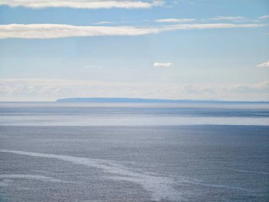 Sakin denizin üzerinde katmanlı bulut şeritleri, ince pastel tonlar ve uzak kara çizgileri ile açık ufuk.