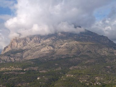 Alçak bulutlarla kaplı dağ, sarp kayalıklarla yükselen yamaç ve desenli granit yüzlü dramatik ışık.