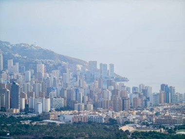 Yoğun kıyı silüeti dağ tabanında kümelenmiş, sisli denizin ötesinde dikey bileşimi oluşturmuş.