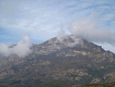 Bulutlarla kaplı puslu dağ zirvesi, engebeli granit yamaçlar ve dokulu kaya yüzeyleri, alçak bulut kümeleri.