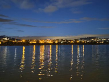 Gece rıhtımında parıldayan şehir ışıkları sakin suyu yansıtıyor, hafif dalgalar dikey ışık yaratıyor.