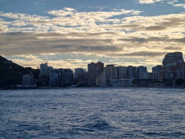 Sabah şehri deniz üzerinde ufuk çizgisi yumuşak altın ışık, durgun su dokuları, siluetli binalar, kayalık