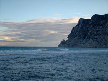 Huysuz uçurum ve huzursuz sörf, düşük ışık dalgaları sarp kayalıklara çarpıyor, ağır bulut bandı