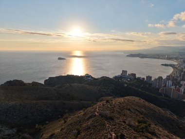 Kıyı kenti üzerinde gün batımı ve kayalıklar altın saatinde deniz yansımasını çerçeveliyor, hava manzarası kenti yakalıyor
