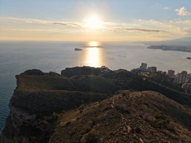 Engebeli tepenin üzerinde altın gün doğumu ve şehrin ufuk çizgisi, güneş sakin okyanusta uzun yansımalar yapıyor.