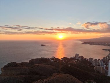 Sıcak turuncu gökyüzü ile koyda canlı günbatımı, son ışığı yakalayan kümelenmiş kuleler, kıvrımlı kıyı şeridi
