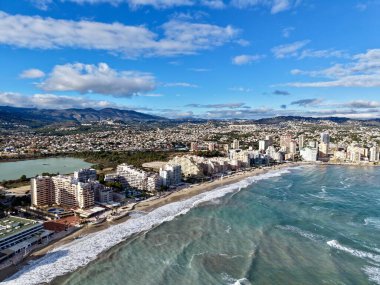 Havada dalgalanan mavi gökyüzü, kumlu sahil kenti, marina boyunca uzanan kuleler.