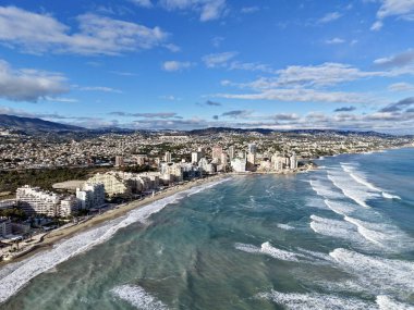 çalkantılı dalgalar ve güneşli binalarla dolu havadan sörf şehri, kalabalık sahil kıyısı boyunca dalgalar çarpışıyor, marina
