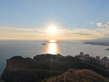 Sahil tepelerinde ve okyanusta gün batımı, sakin sulara yansıyan altın ışık, ufuktaki küçük ada,