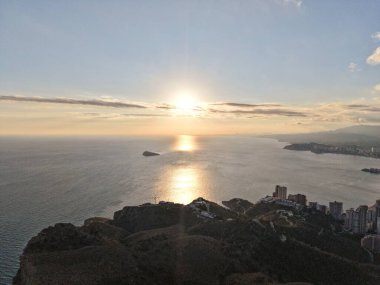 Sahil kenti silueti üzerinde gün batımı yansıması ılık turuncu parıltı ve sakin deniz, alçak bulutlar ve uzak