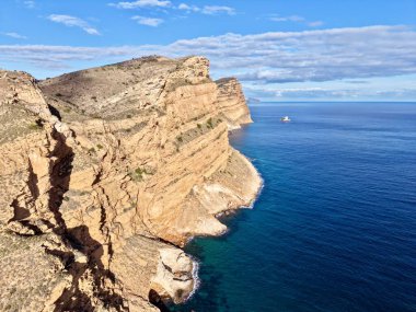 Sıcak, güneşli uçurumların yanında sakin, açık deniz ve uzak tekne, altın tonlarda, parlak katmanlı kaya ve sakin kayalar.