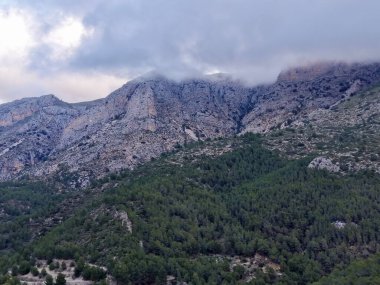 Bulutlarla kaplı engebeli kireçtaşı zirveleri, yoğun kozalaklı yamaçların üzerinde sarp kayalıklar, alçak sis ve kara kara kara kara düşünen