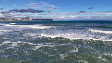 Belirsiz kıyı şeridine doğru ilerleyen uzun dalgalar, sakin deniz ritmi, yumuşak bulutlar, geniş açık ufuk.