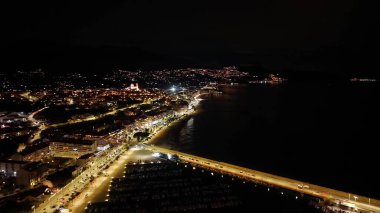 Aydınlatılmış yol ızgarası olan gece limanı kavşağı, karanlık suya uzanan rıhtımlar, şehir kümeleri.