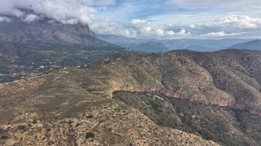 Bulutlu kurak dağ sırtı, engebeli çalı ve dolambaçlı toprak pist, sinematik dron süpürmesi.