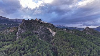 Fırtınalı orman kayalıklarının havadan görünüşü ve kayalıklara tünemiş harabe kaleler, yoğun çam örtüsü ve karamsar bulutlar.