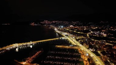 Liman ışıkları sahil limanı marinasını ve köprüyü süpürüyor. Altın parıltı ve deniz manzaralı yollarla aydınlatıyor.