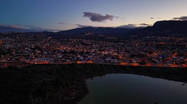 Hava günbatımı şehir ışıkları yamaçtaki panorama yayılan yerleşim bölgesi yansıtıcı suyla, dağla kesişiyor.