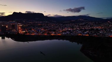 Alacakaranlık Ufuk Çizgisi şehri, ışıklarla parıldayan şehir ışıkları sakin göle yansıyor, dağ silueti çerçevesi.
