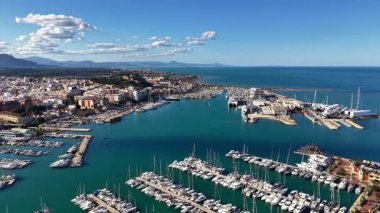 Yatı, turkuaz suyu, güneşli rıhtımı, kalabalık rıhtımı, sahil kenti arka planı, dağ manzaralı marina.