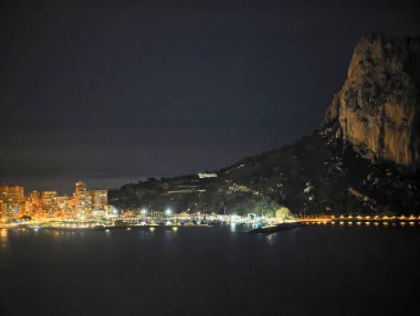 Aydınlatılmış şehir manzaralı gece kıyısı, yumuşak ışıklar izlenen koy, karanlık dağ silueti, sakin deniz