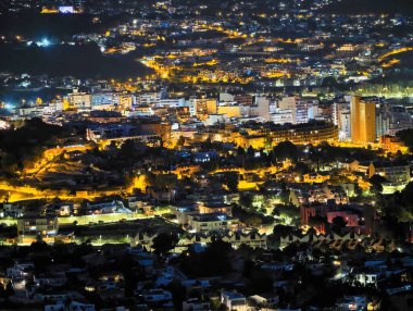 Gece tepe kenti silueti yükseliyor, sıcak altın ışıklar konut yamaçlarına yayılıyor, yoğun kümeler