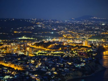 Geceleri geniş kıyı kenti silueti, sokakları ve kıyı şeritlerini, uzak tepeleri izleyen geniş açık kurdeleler.