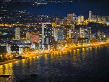 Geceleri deniz önü silueti, durgun suyu, gezinti ışıklarını ve canlı ışığı yansıtan parlak gökdelenler.