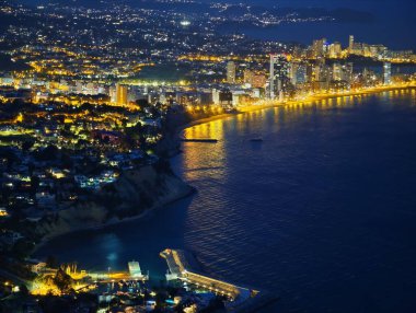 Gezinti ışıkları denizi kucaklıyor, sahil kuleleri kehribar ışığı yansıtıyor, nazik dalgalar öpüşüyor.