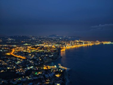 Aydınlatılmış kıyı şeritleri ve kümelenmiş mahalleleriyle dramatik gece kıyısı eğrisi, geniş karanlık deniz
