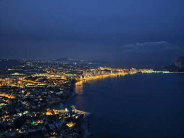 Kıvrımlı kıyı şeridi alacakaranlıkta aydınlandı. Altın sokak lambaları sahili kucakladı, binalar istiflendi.