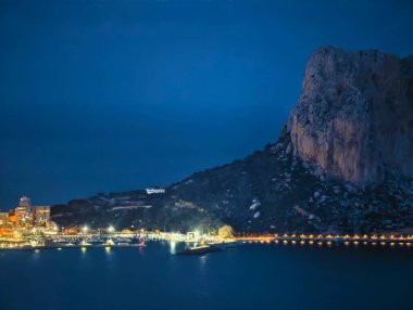 Aydınlatılmış temeli olan gece kayalıkları sakin okyanusun üzerindeki engebeli kaya yüzeyi, yol ışıkları...