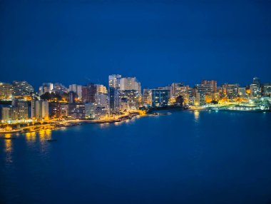 Neon ve ılık tonlarda aydınlanan modern gökdelenlerle dolu canlı deniz manzaralı gökyüzü, sakin koy yansıtan kuleler.