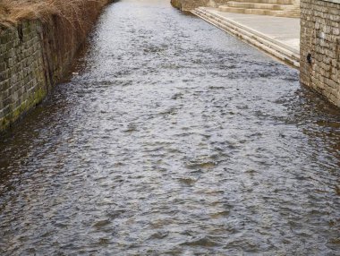 Buz gibi suyu akan taş kanallar, çakıl taşlı bankalar ve yosunlu duvarlar, sağda dar geçit, bulutlu