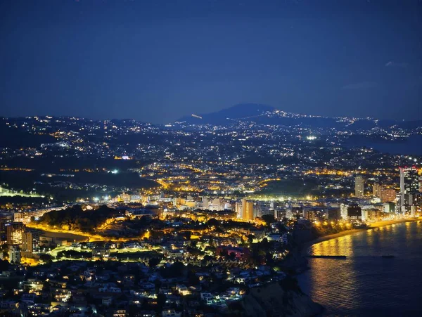 Gece kıyısı kıyısı kıyı şeridi kıyı şeridi boyunca parıldayan yansımalar yoğun kentsel ışıklar, sakin deniz ve uzak mesafeler.