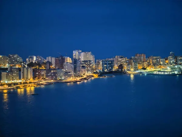 Neon ve ılık tonlarda aydınlanan modern gökdelenlerle dolu canlı deniz manzaralı gökyüzü, sakin koy yansıtan kuleler.