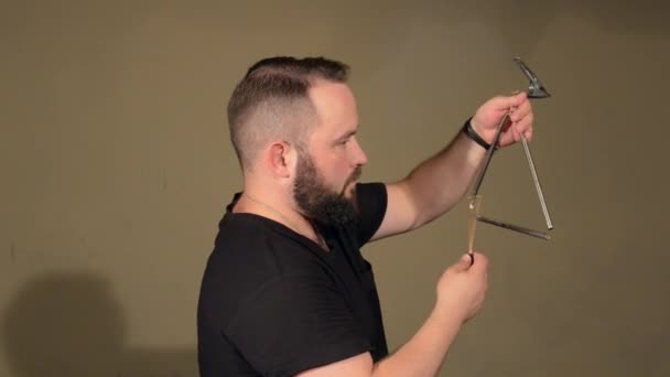 En man med skägg i svart T-shirt, spelar på slagverk instrument triangeln. på de ljusa väggarna bakgrund. — Stockvideo