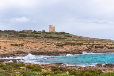 Tarihi kare bir taş gözetleme kulesi, kayalık bir kıyı şeridine bakan çimenli bir tepenin üzerinde ve bulutlu bir gökyüzünün altında dalgaları olan turkuaz Akdeniz 'de bulunur..